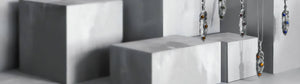 La imagen muestra varios collares en forma de pluma de plata con pequeñas piedras de colores incrustadas, suspendidos sobre bloques geométricos de tonos grises.