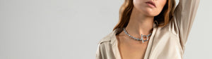 La imagen muestra a una mujer vistiendo una camisa de seda beige, luciendo un collar grueso de plata con un cierre frontal en forma de omega, posando con el brazo levantado.