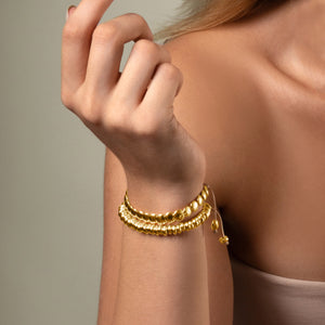 Mujer con pulsera dorada de cuentas múltiples en la muñeca, con fondo neutro y el brazo parcialmente levantado.