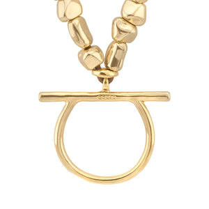 Detalle del colgante central de un collar dorado con cuentas irregulares, que muestra un aro grande unido a una barra horizontal con la marca 'Corso' grabada, destacando el acabado brillante y el diseño moderno de la pieza.