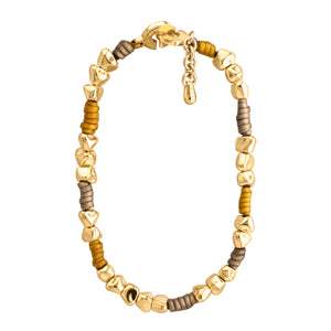 Pulsera de diseño moderno que combina cuentas doradas irregulares con secciones de cuero color taupe y mostaza. Las cuentas doradas tienen formas únicas y facetadas, dando un aspecto orgánico y artesanal. El cuero color taupe y mostaza está dispuesto en pequeñas secciones entre grupos de cuentas doradas. La pulsera tiene un cierre circular dorado con un diseño distintivo y grabado con el texto "CORSO". Una pequeña cadena dorada cuelga del cierre, probablemente para ajustar el tamaño. El contraste entre el o