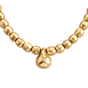 Primer plano de un collar dorado elegante. Está compuesto por cuentas esféricas de oro pulido, ligeramente irregulares en su forma, que le dan un aspecto artesanal y orgánico. En el centro del collar cuelga un dije en forma de gota, también de oro, más grande que las cuentas. El collar tiene un brillo suave y cálido, característico del oro. El fondo es blanco, lo que hace resaltar los detalles y el color del collar.