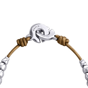 Detalle del cierre de un collar o pulsera de joyería. El cierre es una pieza circular plateada con un diseño de gancho, que tiene grabado 'corso' en su superficie. Este cierre dorado está conectado a un cordón de cuero marrón en ambos lados, que está anudado alrededor del cierre. A cada lado del cordón de cuero se ven algunas cuentas plateadas, probablemente del resto de la joya. El fondo de la imagen es blanco, lo que hace resaltar los detalles del cierre y los materiales.