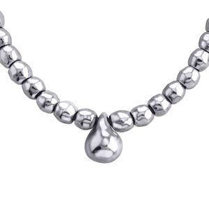 Primer plano de un collar plateado elegante. Está compuesto por cuentas esféricas de plateadas, ligeramente irregulares en su forma, que le dan un aspecto artesanal y orgánico. En el centro del collar cuelga un dije en forma de gota, también plateado, más grande que las cuentas. El collar tiene un brillo suave y cálido, característico de la plata. El fondo es blanco, lo que hace resaltar los detalles y el color del collar.