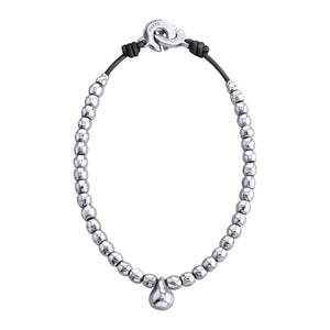 Una imagen de un collar elegante compuesto por esferas plateadas uniformes, enhebradas en un cordón negro. En el centro del collar cuelga un pequeño dije o colgante también plateado con forma redondeada. El cierre del collar es una pieza circular plateado con un diseño distintivo, conectada al cordón negro en ambos extremos. Este collar combina el brillo del metal precioso con la textura oscura del cordón, creando un contraste atractivo y sofisticado.
