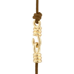 Esta imagen muestra un detalle de una joya o collar. Se ve un cordón de cuero marrón del que cuelga un gancho o cierre dorado. El gancho dorado tiene un diseño decorativo con pequeñas ondulaciones o anillos en ambos extremos. La parte superior del gancho está unida al cordón de cuero mediante un nudo, mientras que la parte inferior tiene un aro abierto, presumiblemente para enganchar la otra parte del collar. El contraste entre el cuero marrón y el oro brillante crea un aspecto elegante y artesanal.