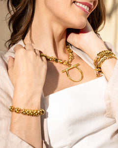 La imagen muestra a una mujer sonriente luciendo joyas doradas. Lleva un collar dorado con un colgante en forma de U o herradura y pulseras doradas en ambas muñecas. Viste una blusa blanca y una chaqueta clara. Se enfoca en el cuello, pecho y manos de la mujer, mostrando de cerca las joyas. Su sonrisa y cabello castaño son visibles parcialmente.