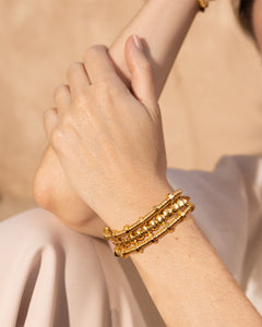 Mano con pulsera dorada de múltiples hilos y cuentas, sobre fondo beige claro.
