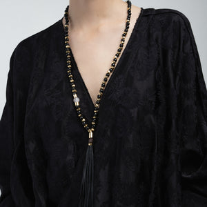 Primer plano de un collar de cuentas negras y doradas con una borla negra, usado sobre una blusa negra de textura floral.