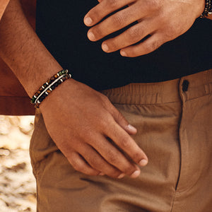 Primer plano de una muñeca masculina luciendo varias pulseras de cuentas en tonos marrones, verdes y negros, complementando un atuendo casual con pantalones beige.